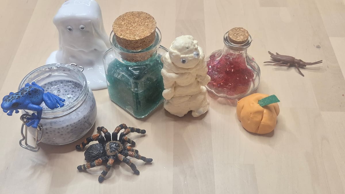 Six homemade slimes on a wooden table: a white slime ghost, chia frog spawn with a blue toy frog, green monster snot in a jar, a marshmallow mummy, red jelly goo in a bottle and an orange slime pumpkin, guarded by a plastic tarantula